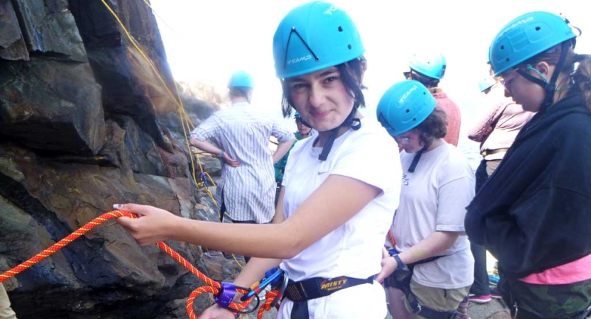 A student in a blue helmet and other safety gear looks at the camera and holds a rope. There are others. behind them. 